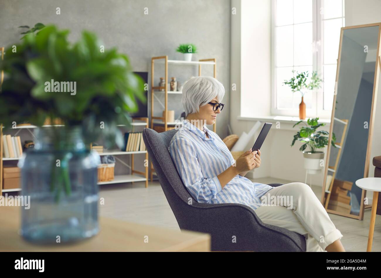 Relaxed senior woman sitting in armchair at home and reading book on digital tablet Stock Photo