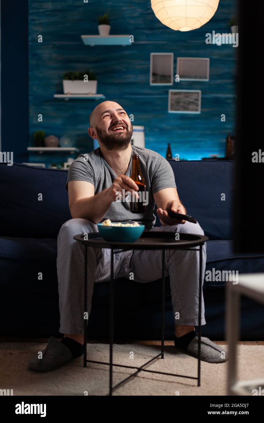 Excited relaxed man spending free time watching TV entertainment series eating popcorn and drinking beer. Funny amused enjoying evening at home sitting on comfortable couch dressed in pajamas Stock Photo
