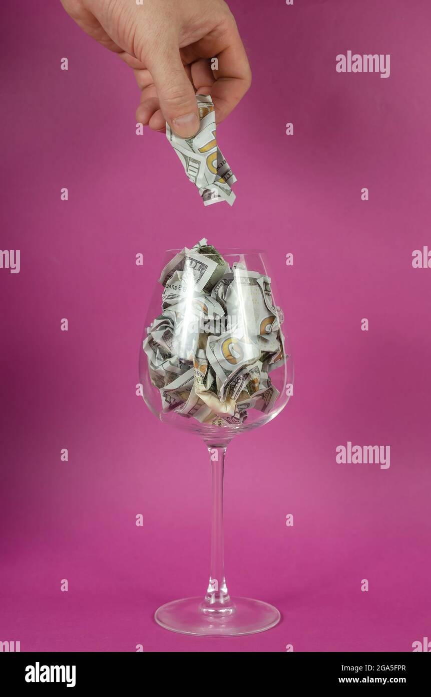 Male hand puts money in wine glass against pink background. Glass is full of crumpled hundred dollar bills. Stock Photo