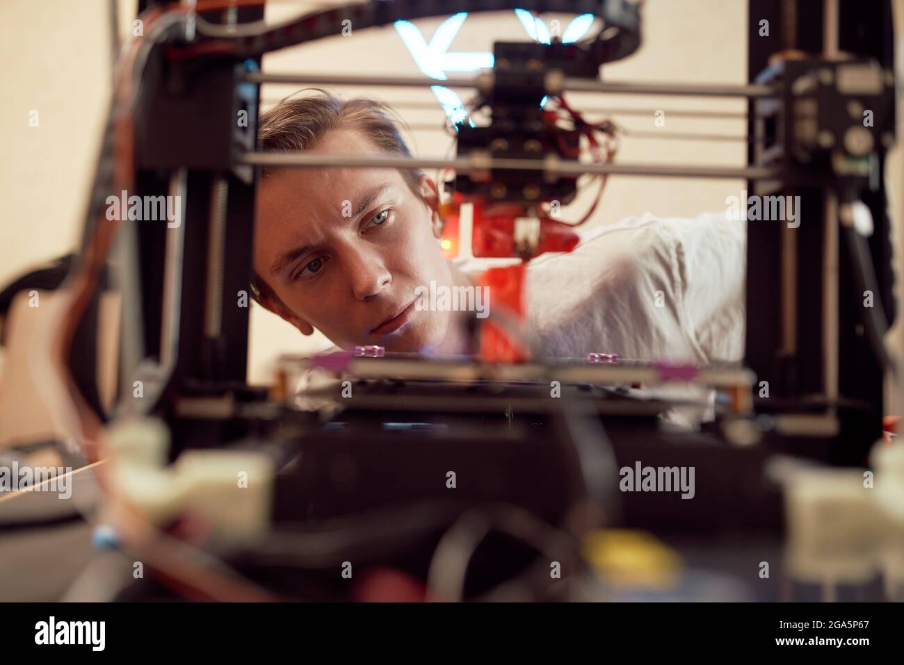 Focused young man watching modern 3D printer creating red plastic model in studio Stock Photo