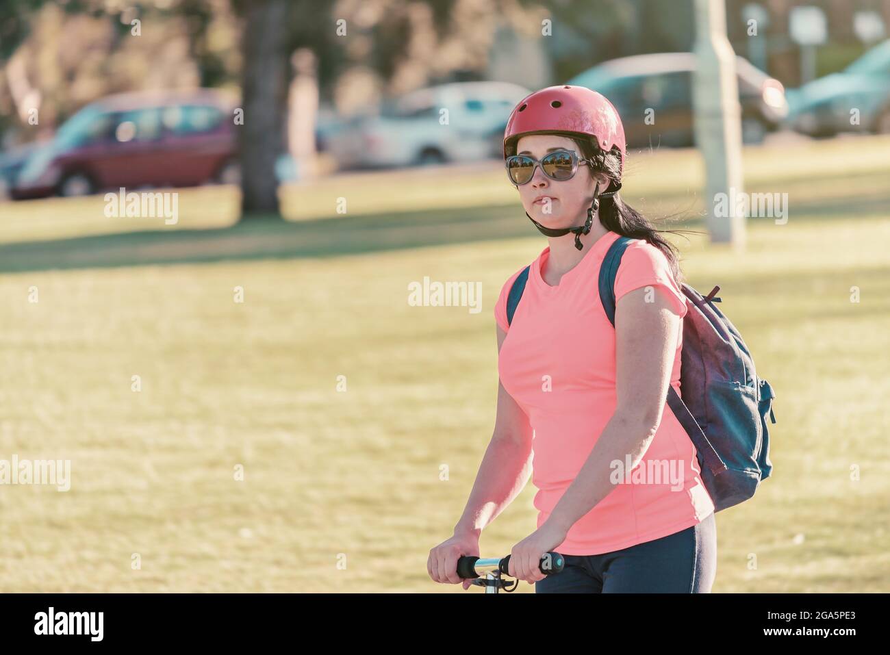 Portrait of young woman riding push scooter through Adelaide Parklands while  wearing sunglasses with pink top and bike helmet Stock Photo