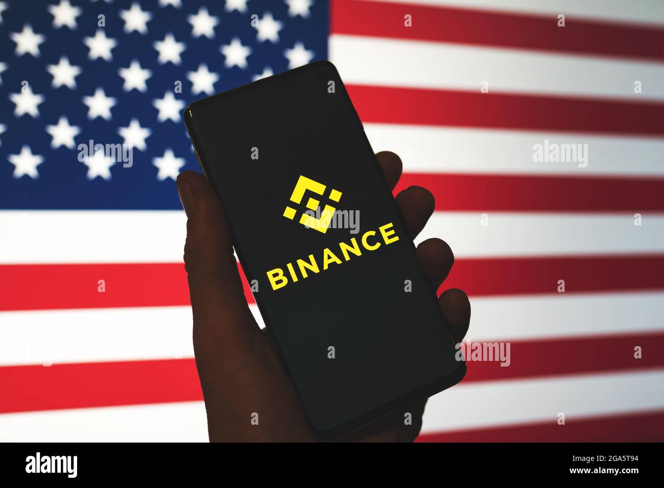 Binance app logo on smartphone in hand with blurred American flag background. Binance in the USA news. Crypto exchange, trading platform. Swansea, UK - July 27, 2021. Stock Photo