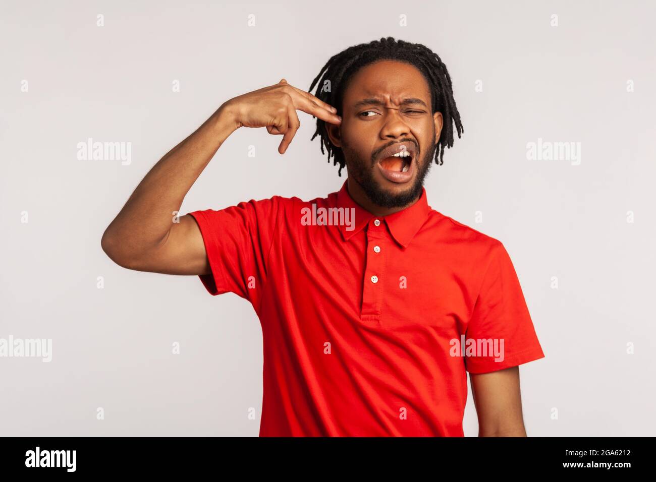 Bearded man wearing red casual style T-shirt, holding fingers near temple pretending holding gun, going to make shot, faced with problems and crisis. Stock Photo