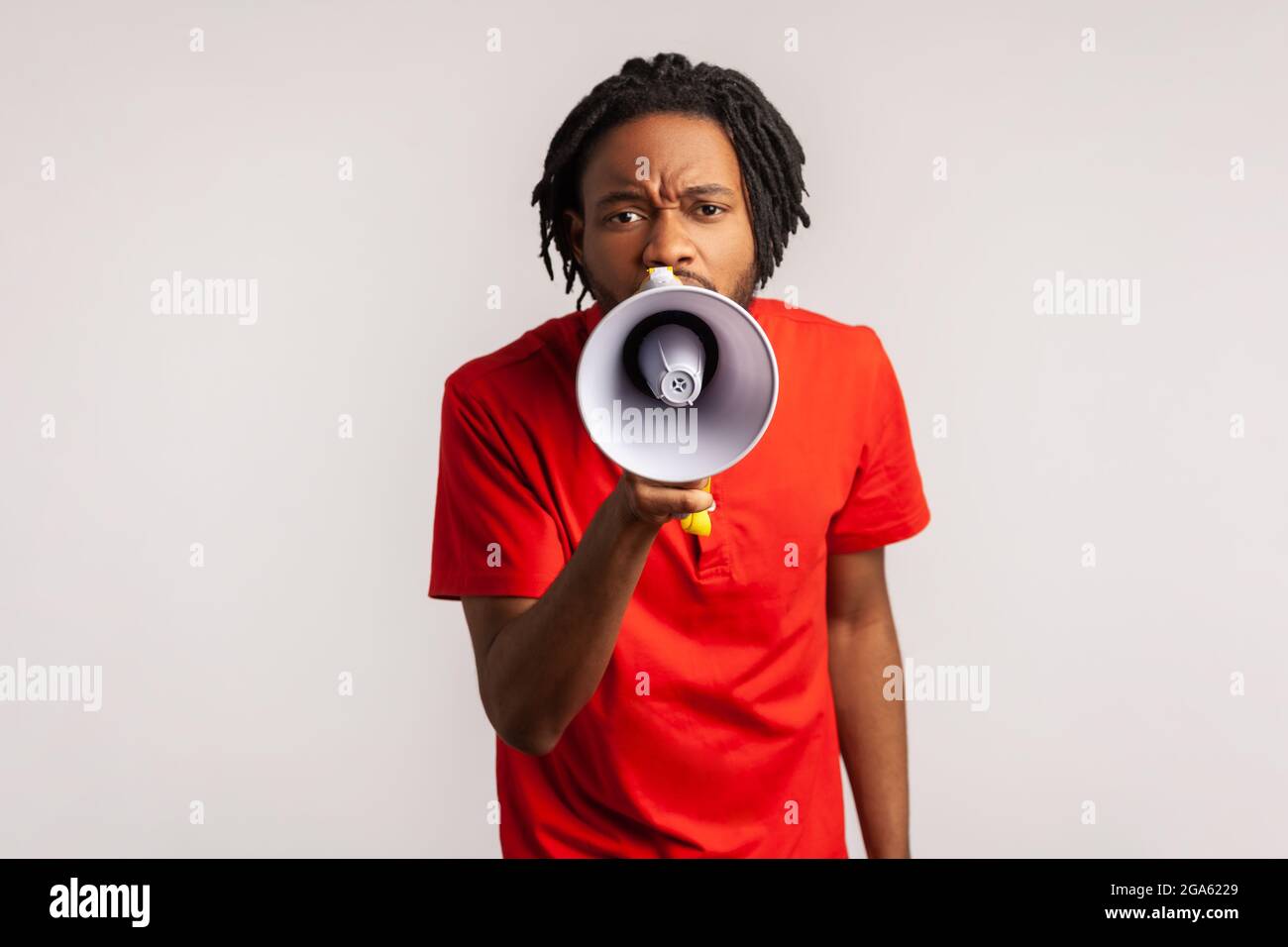 Man with dreadlocks wearing red casual style T-shirt, holding megaphone near mouth loudly speaking, screaming, making announcement, paying attention. Stock Photo