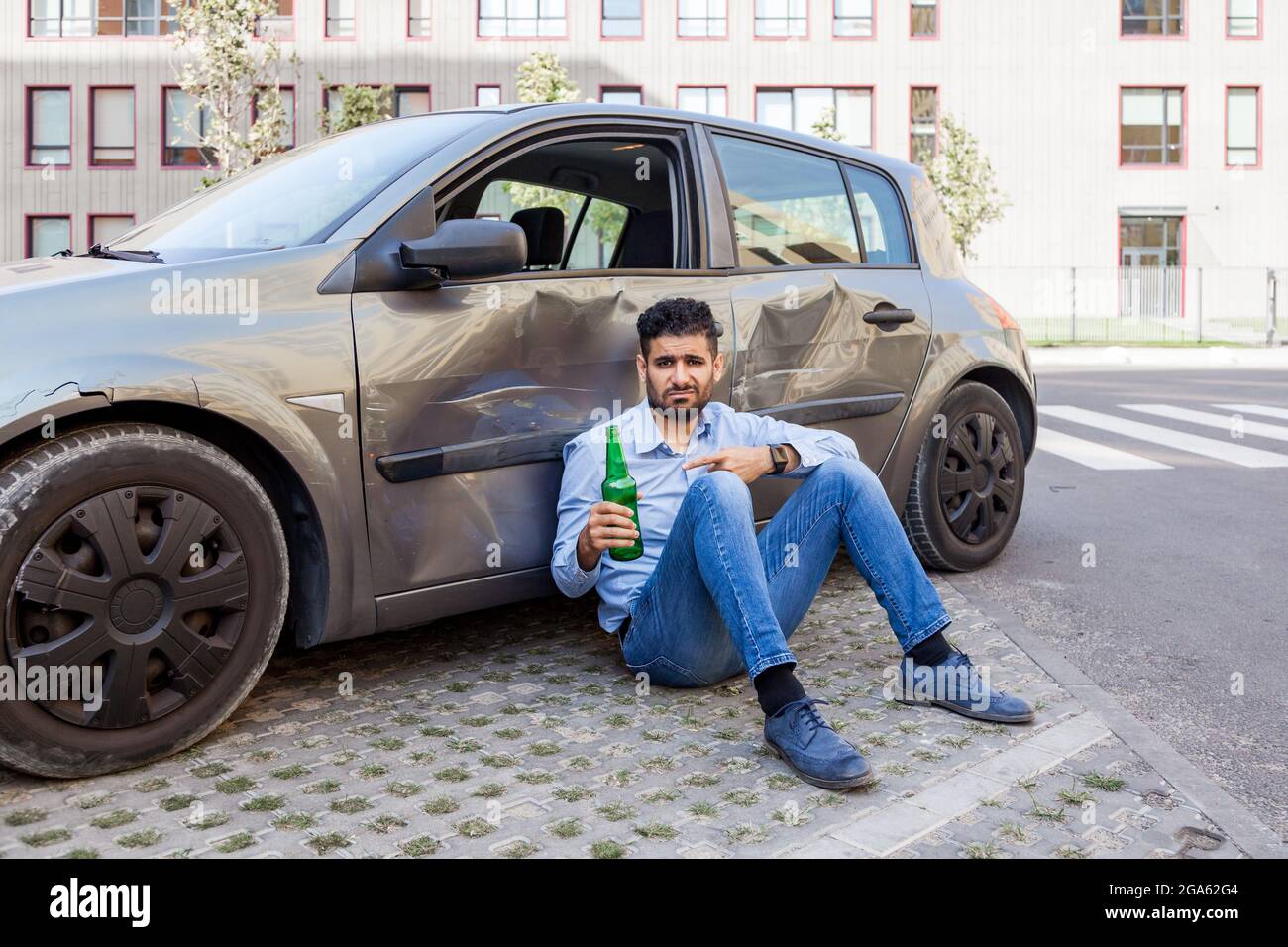 Upset bearded man wearing blue shirt and jeans sitting near smashed car