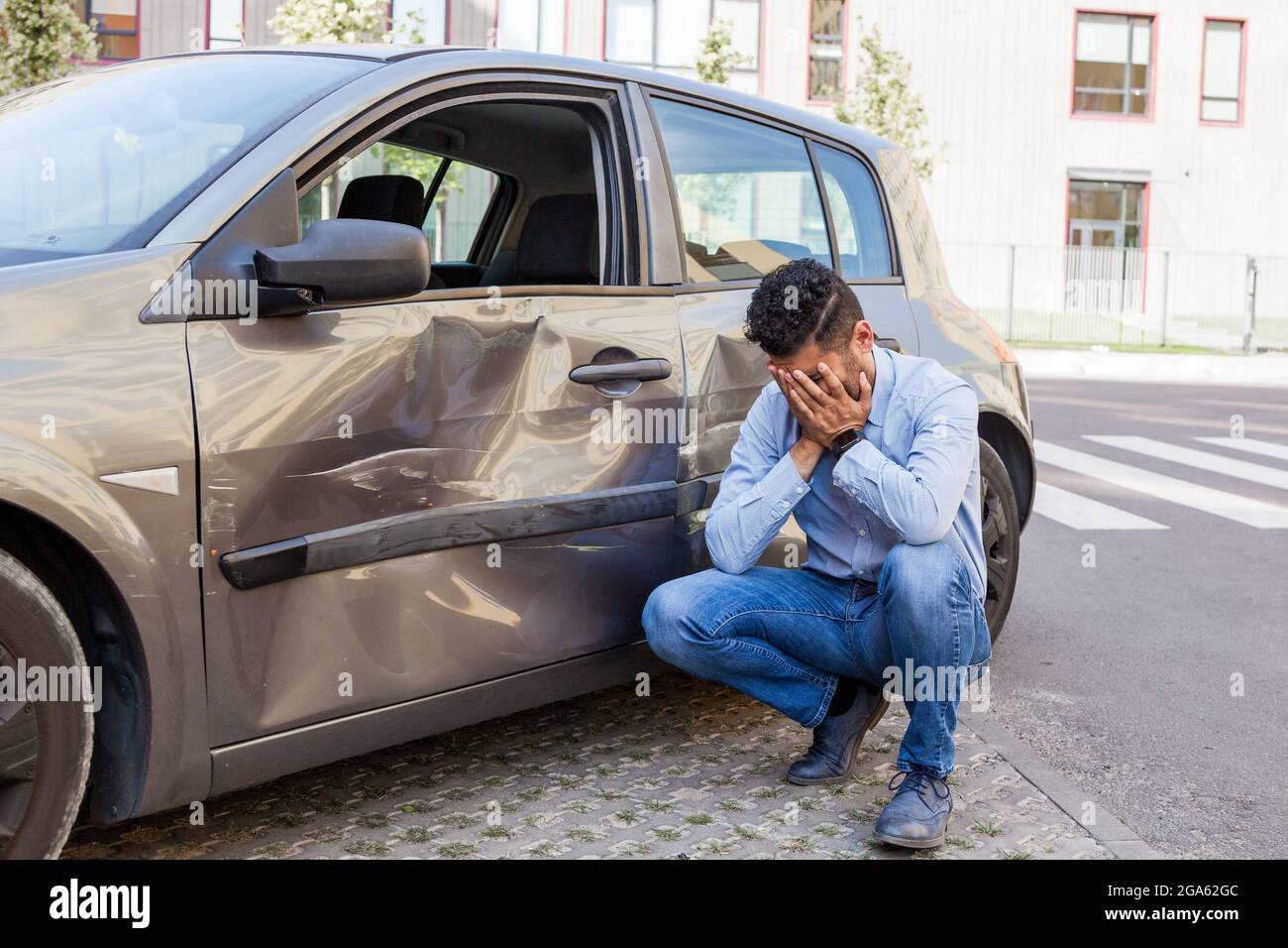 Portrait of man wearing jeans and blue shirt broken his car, being upset and covering his face with palms, dents and scratches on the door of auto, da Stock Photo