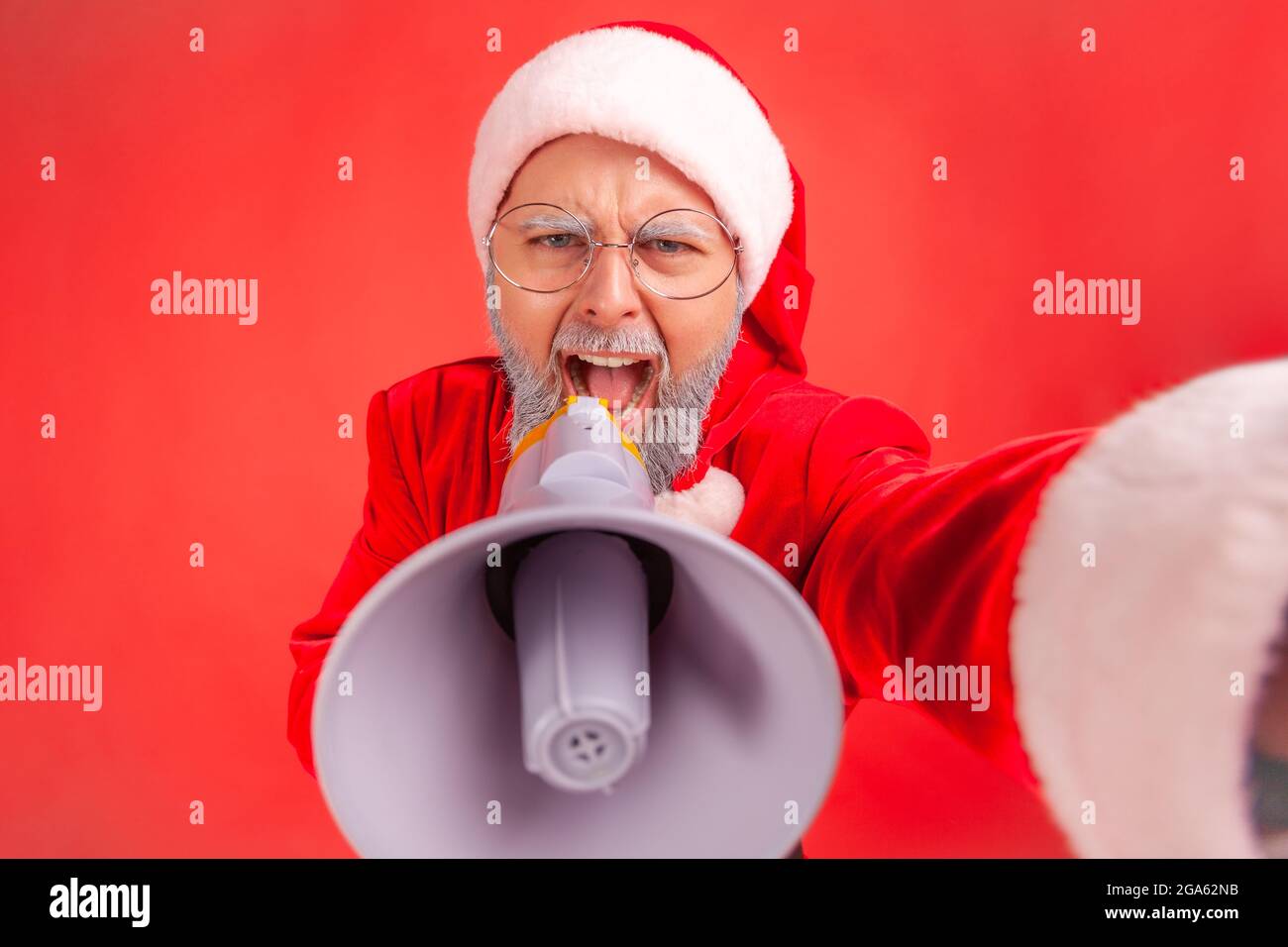 Crazy elderly man with gray beard wearing santa claus costume screaming at megaphone, protesting while livestreaming, wants to be heard POV. Indoor st Stock Photo