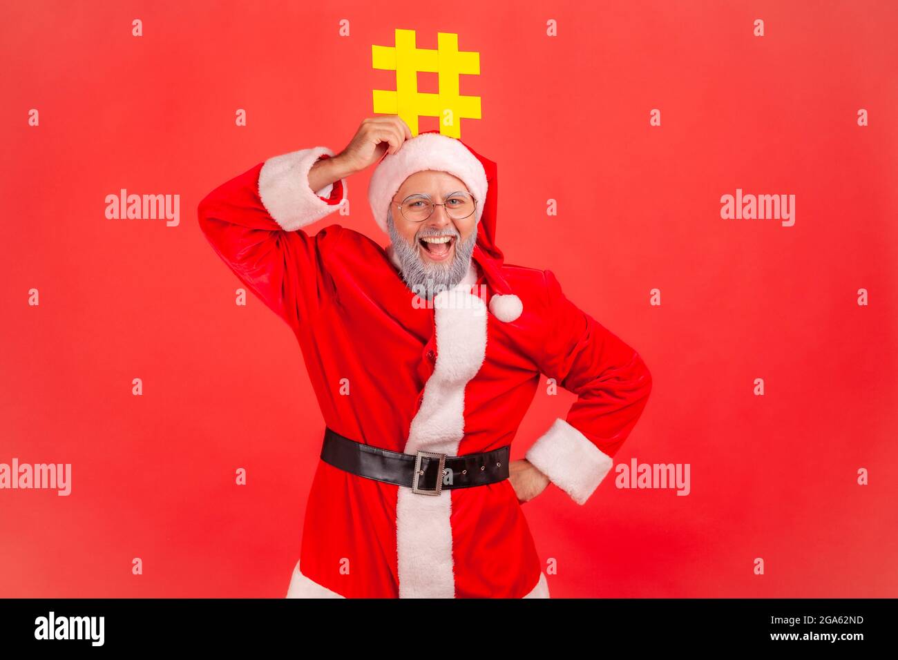 Smiling elderly man with gray beard wearing santa claus costume holding hashtag symbol over head, promoting viral topic, trendy idea, celebrating. Ind Stock Photo