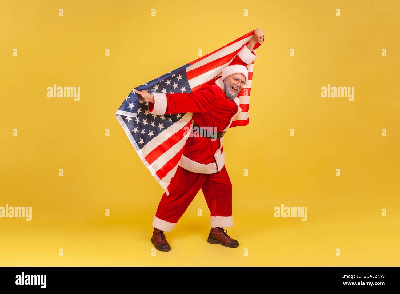 Smiling elderly man with gray beard wearing santa claus costume holding USA flag in hands, looking at camera, pretending flying, celebrating new year. Stock Photo