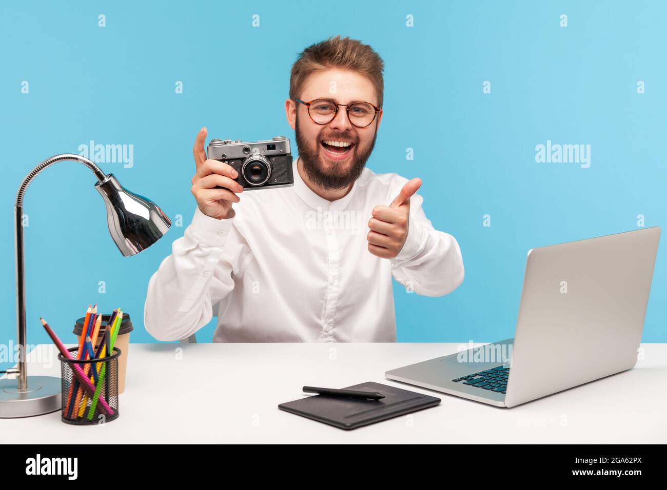 Cheerful excited cameraman showing thumbs up holding vintage photocamera and smiling looking at camera, sitting at workplace, satisfied with photo sho Stock Photo