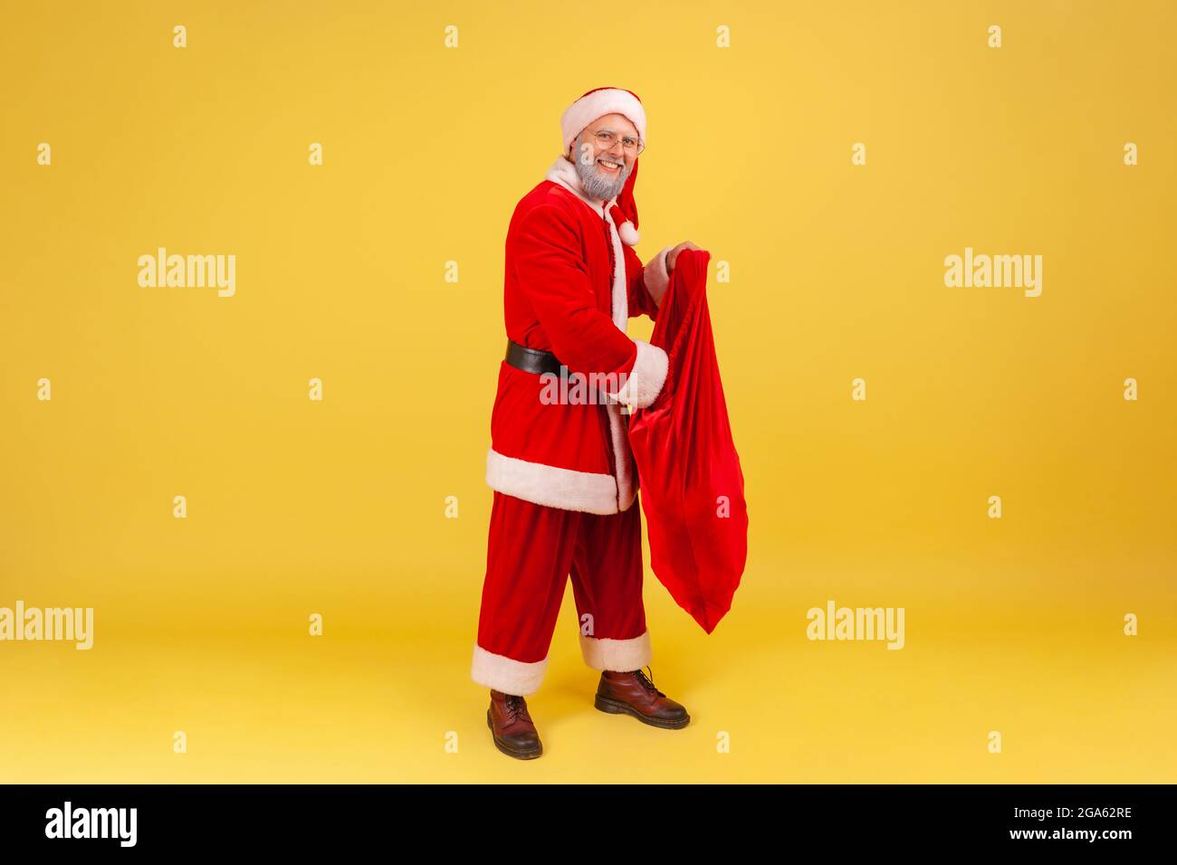 Smiling elderly man with gray beard wearing santa claus costume putting hand inside big red bag full of presents for Christmas, greeting with new year Stock Photo