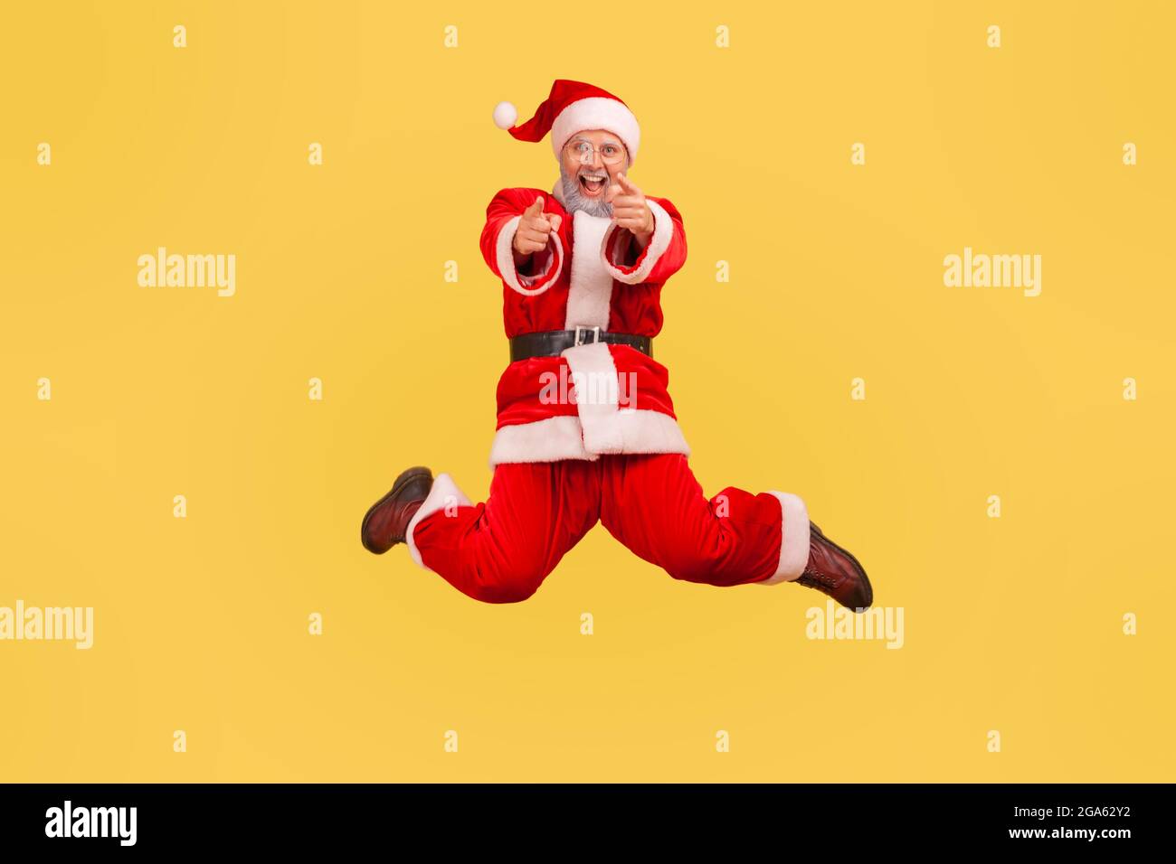 Elderly man with gray beard wearing santa claus costume jumping high and looking at camera, pointing with fingers, satisfied with winter holidays. Ind Stock Photo