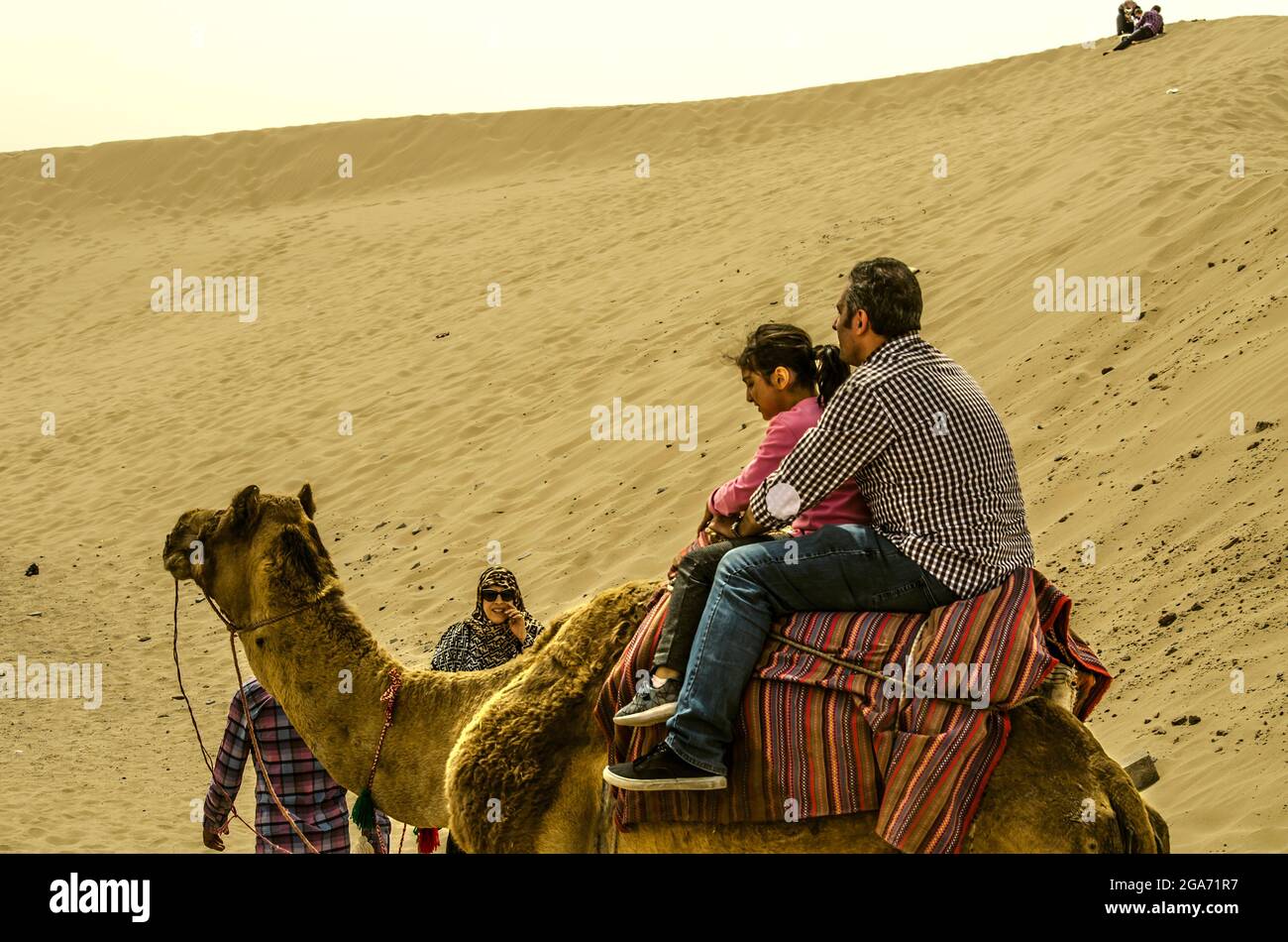 Yazd, Desert, Iran, February 20, 2021: Man with a little girl sitting on a camel covered with a bright carpet, accompanied by a guide and a woman watc Stock Photo