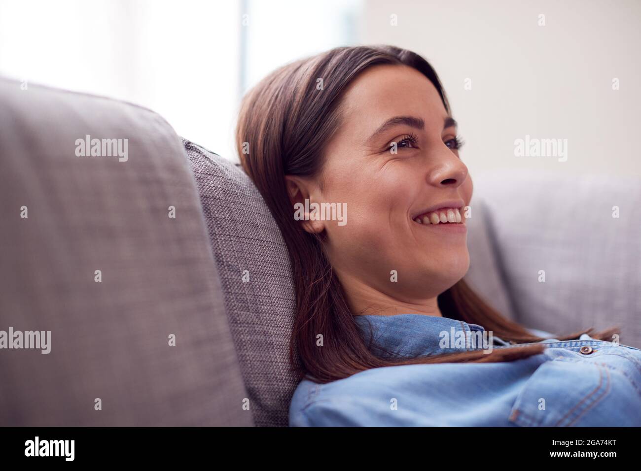 Smiling Young Woman At Home Lying On Sofa Watching TV And Laughing Stock Photo