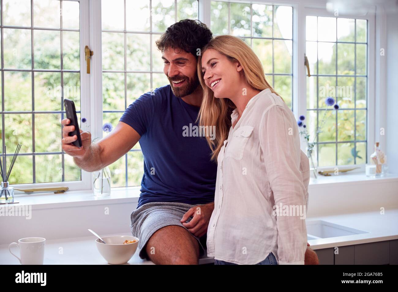 Couple Wearing Pyjamas In Kitchen Using Mobile Phone Whilst Eating Breakfast Stock Photo