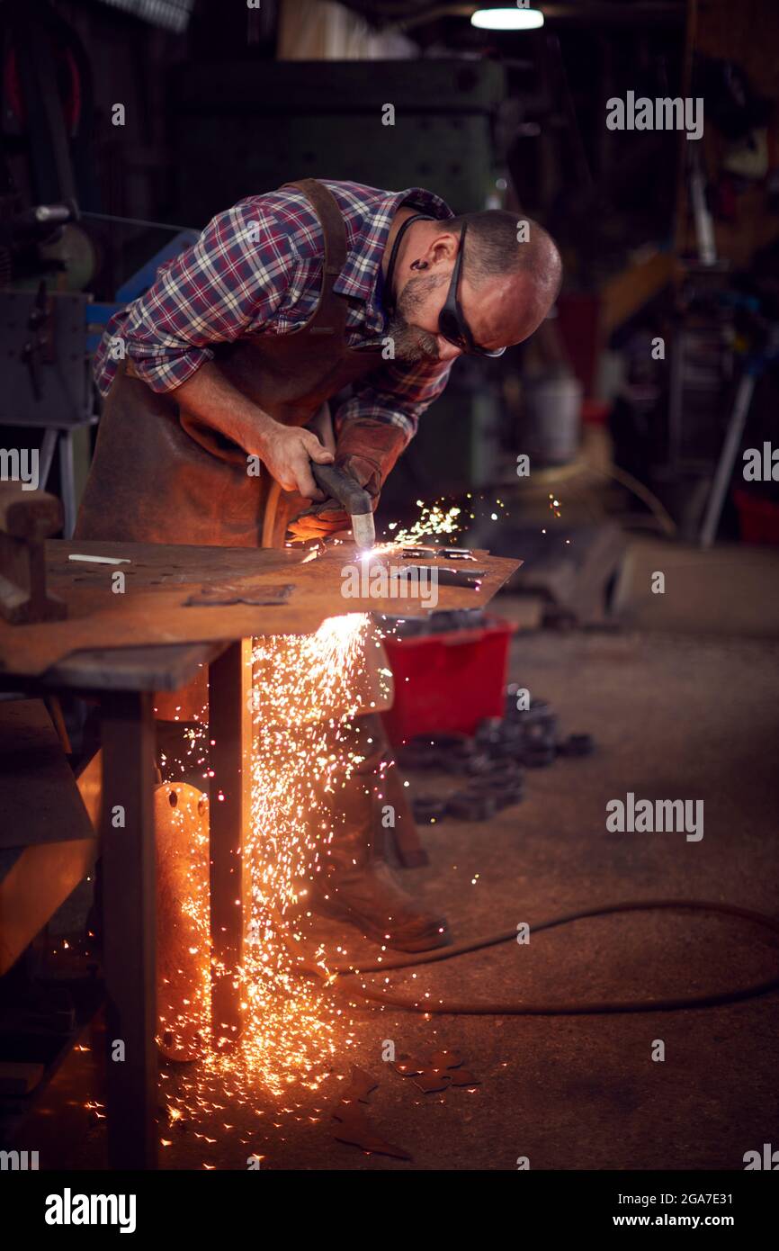 Male Blacksmith Using Plasma Cutter To Cut Shape From Sheet Metal In Forge Stock Photo