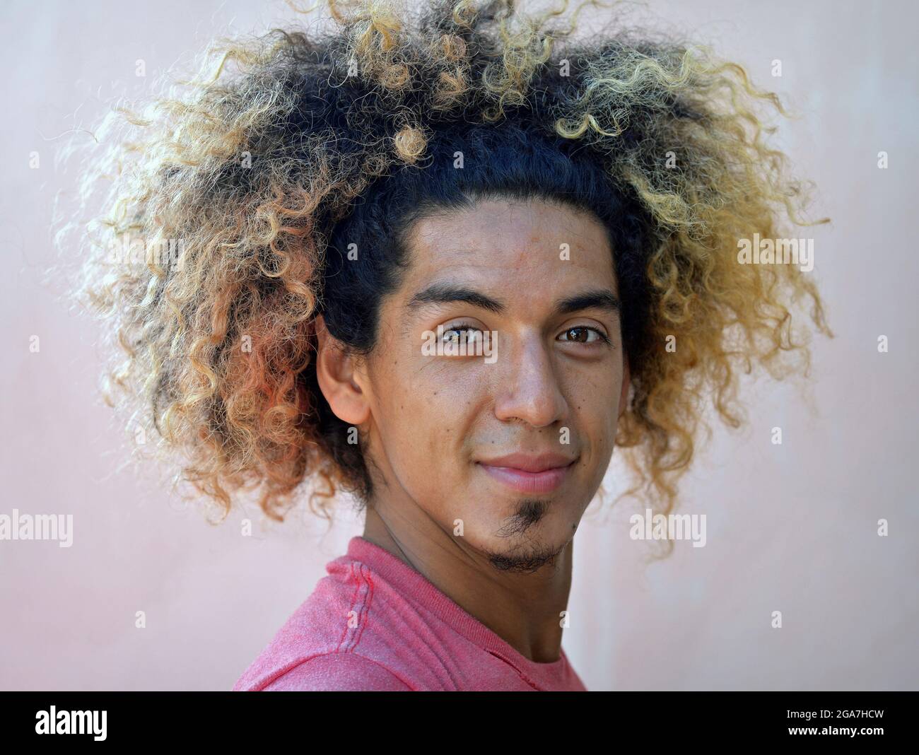 Young Latin American man with a shock of wild curly hair and blond dyed hair ends smiles with closed mouth at the camera. Stock Photo