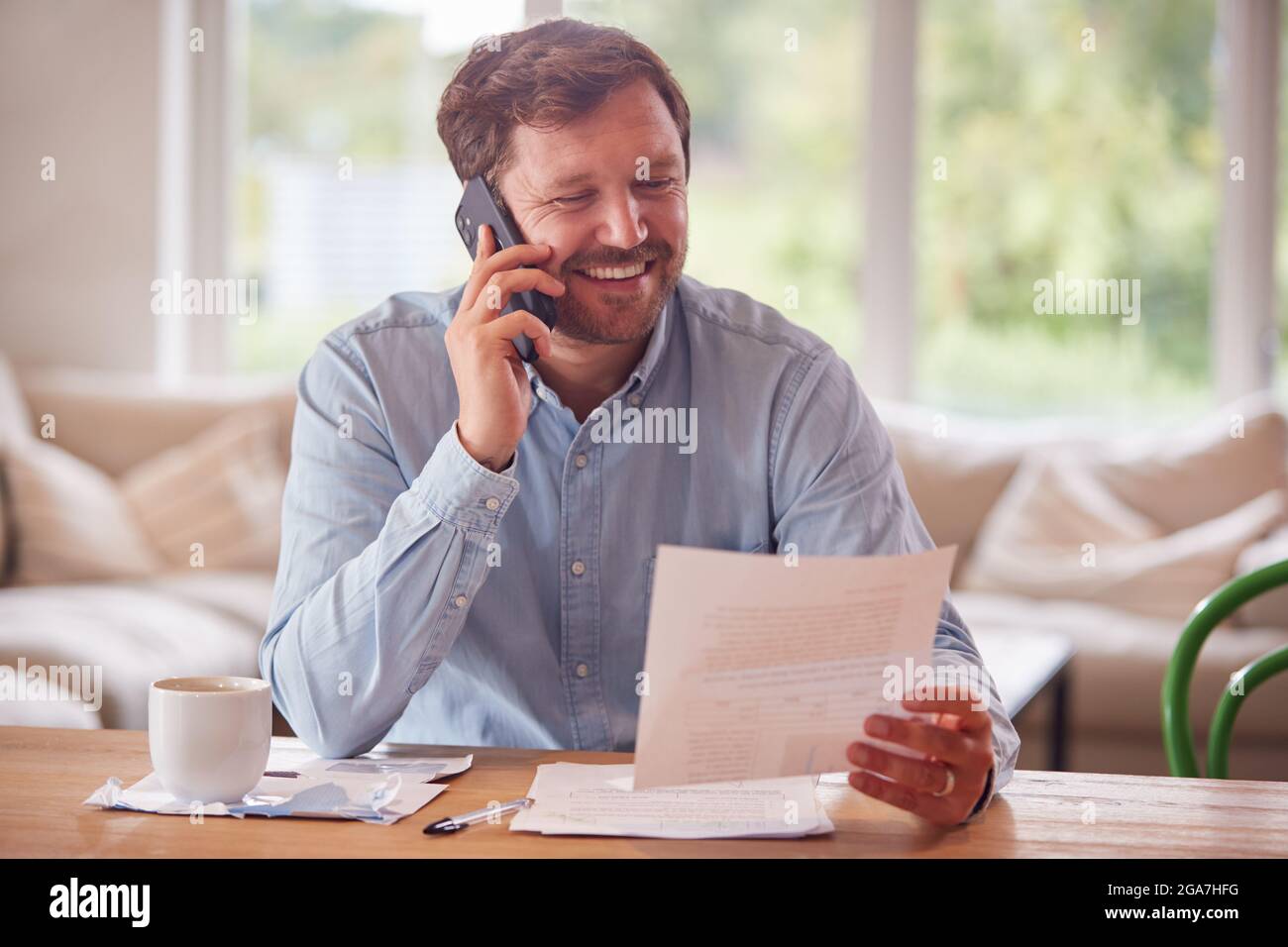 Smiling Man On Phone Call Sitting At Table At Home Reviewing Domestic Finances Stock Photo
