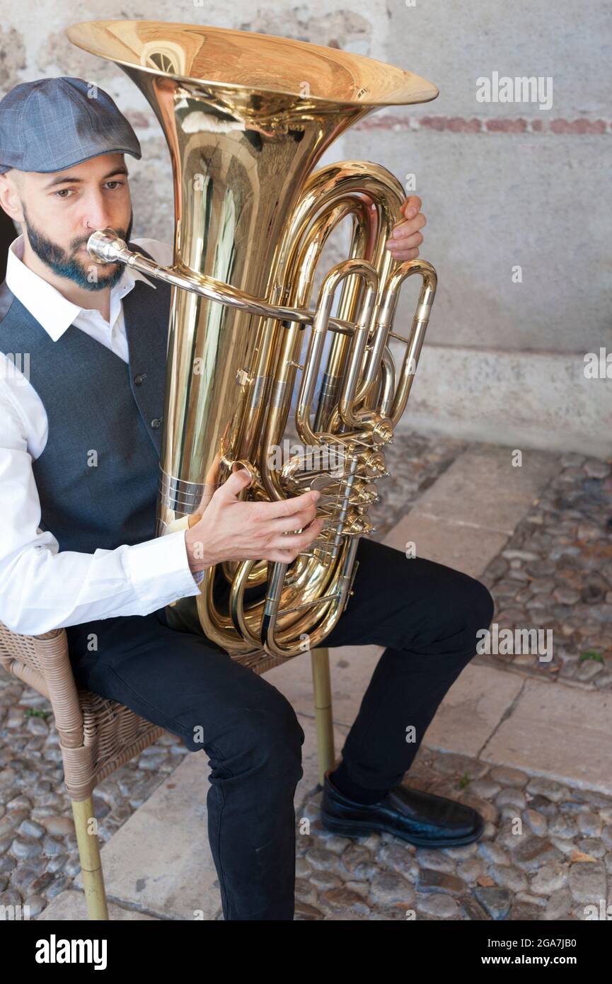 A jazz musician dressed in vintage style, playing the tuba on the street in broad daylight. With beard and goatee dyed blue and a retro cap. Stock Photo