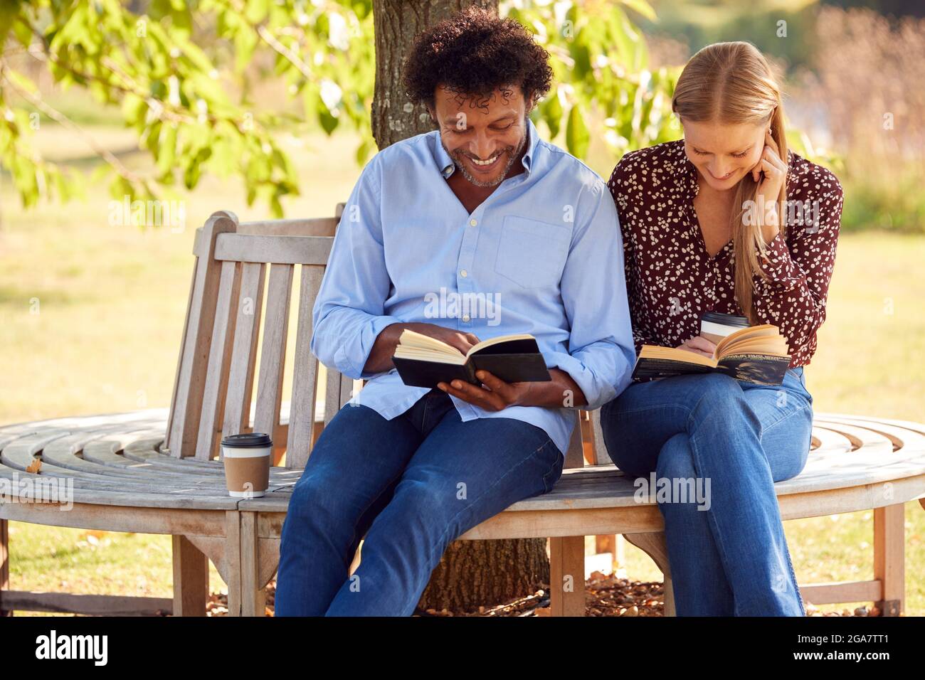 Mature Couple Relaxing Sitting On Park Bench Under Tree Reading Books With Takeaway Coffees Stock Photo