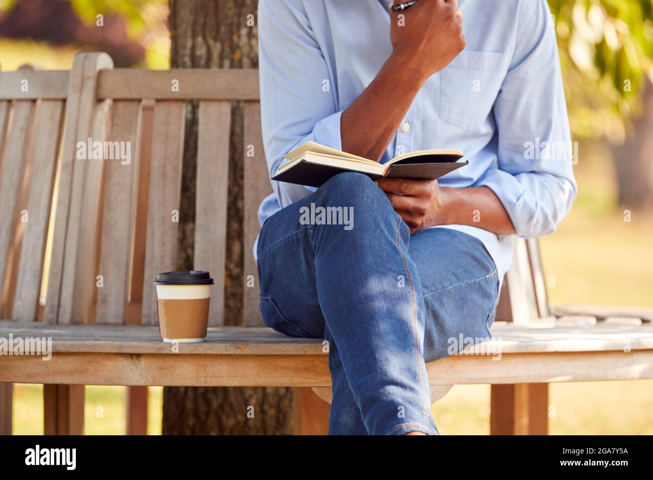 Close Up Of Man With Takeaway Coffee Sitting On Park Bench Under Tree Writing In Notebook Stock Photo
