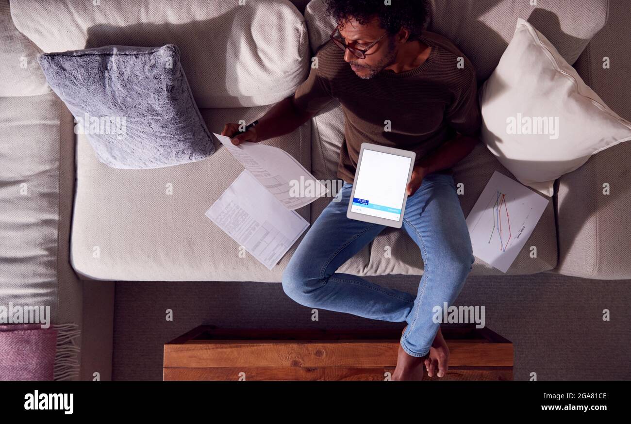 Overhead Shot Of Man Working From Home Sitting On Sofa Reviewing Documents Using Digital Tablet Stock Photo