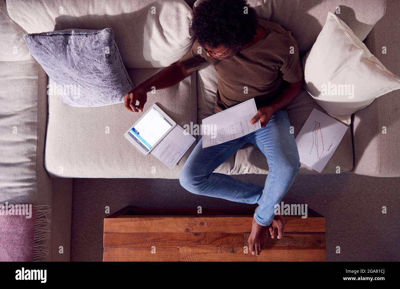 Overhead Shot Of Man Working From Home Sitting On Sofa Reviewing Documents Using Digital Tablet Stock Photo