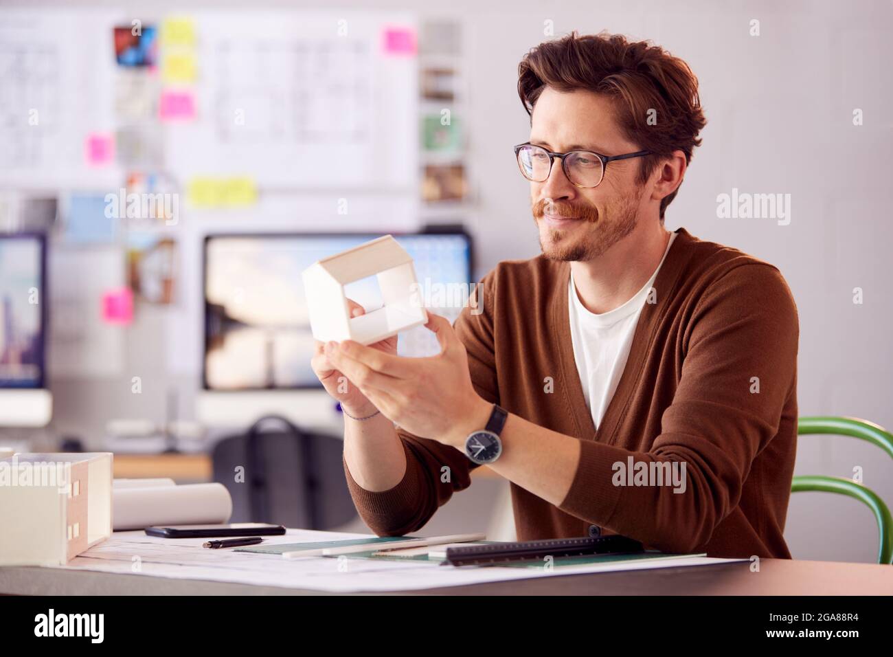 Male Architect In Office Working At Desk With Wooden Model Of Building Stock Photo