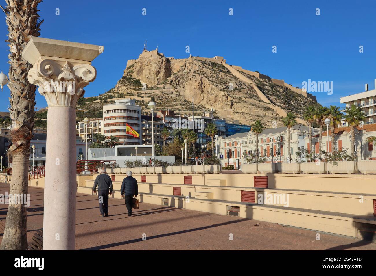The castle of Santa Barbara high up on a promontory overlooking the city of Alicante, Spain with the esplanade and people walking in the foreground Stock Photo