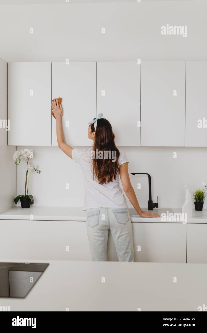 Rear view of young woman cleaning surface of white kitchen wall cabinet with rag and spray bottle detergent. Stock Photo