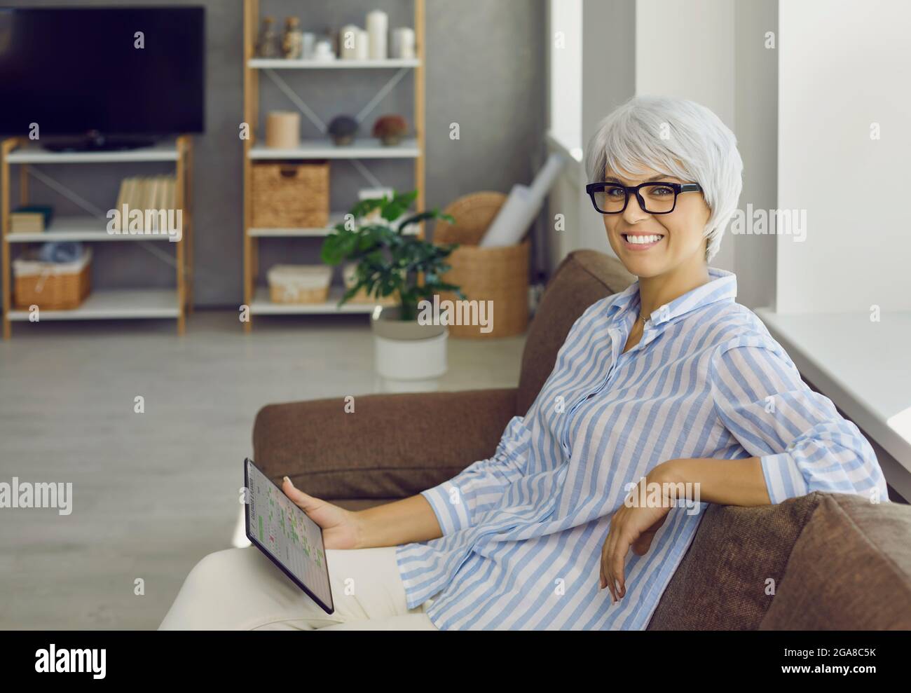 Happy mature woman sitting on sofa and planning her time using planner app on tablet Stock Photo