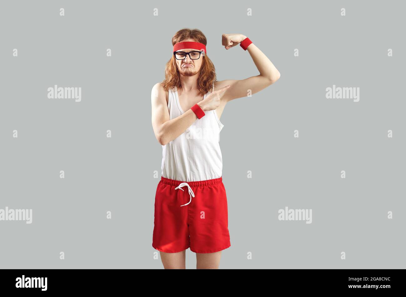 Funny slim guy stands on a gray background and shows his small biceps, which he is not happy with. Stock Photo