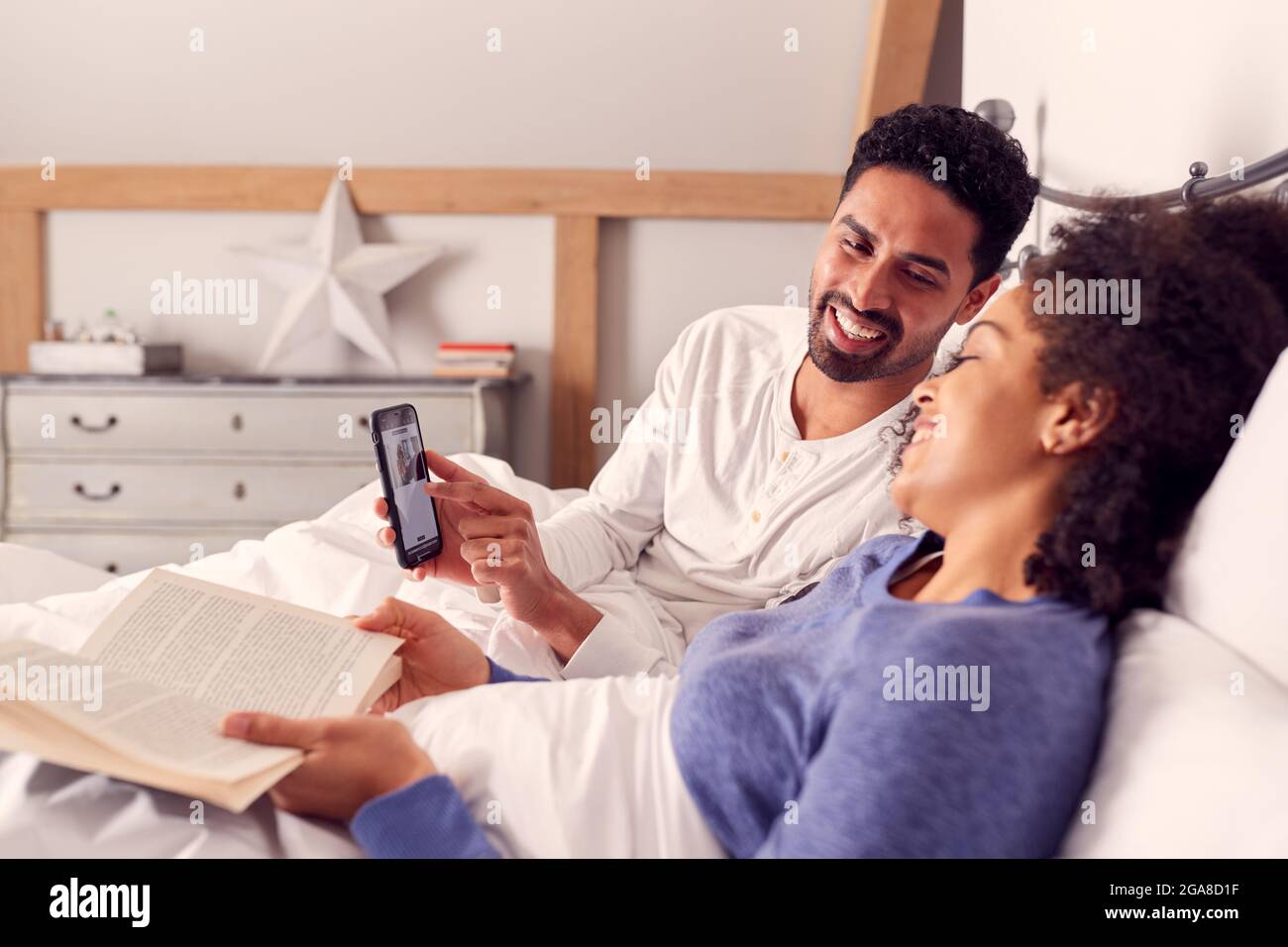 Couple In Bed Reading And Looking At Photos On Mobile Phone Together Stock Photo