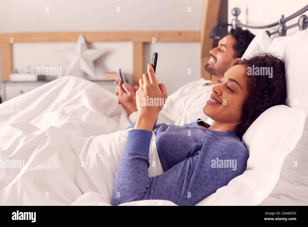Couple In Wearing Pyjamas Lying In Bed Looking At Mobile Phones Together Stock Photo