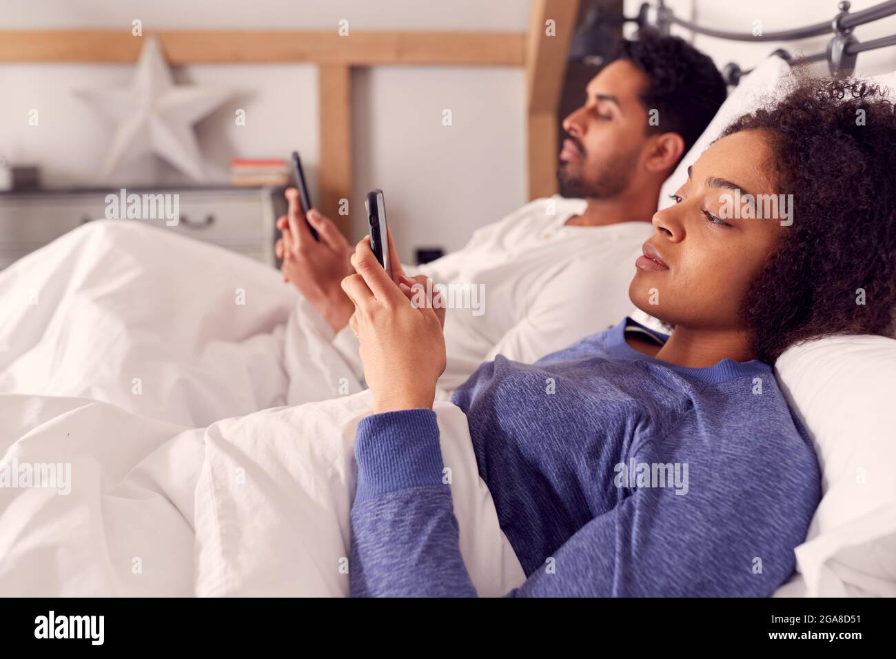 Couple In Wearing Pyjamas Addicted To Using Mobile Phones Lying In Bed And Not Communicating Stock Photo