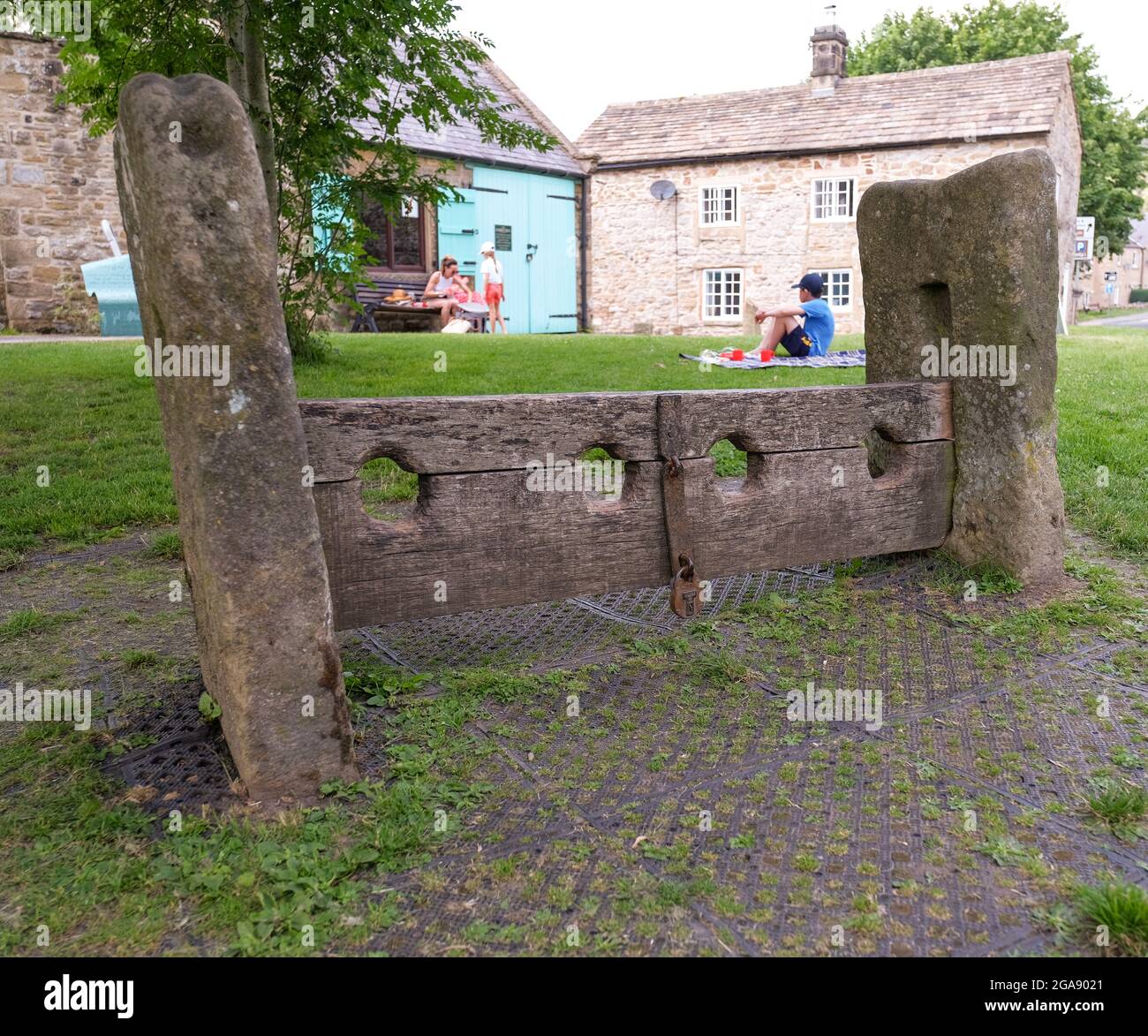 The wooden stocks in the Derbyshire plague village of Eyam. In the background are 2 children playing on the green enclosed by 2 stone buildings. Stock Photo