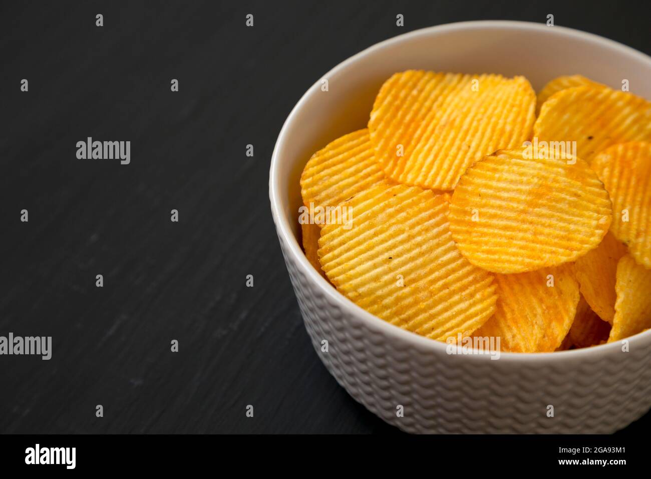 Ruffled Cheese Potato Chips in a Bowl on a black surface, side view. Space for text. Stock Photo