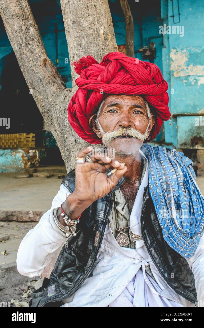 Portrait of Indian male villager in a red turban and tribal jewelry smoking a cigarette in a village in Rajasthan, India Stock Photo