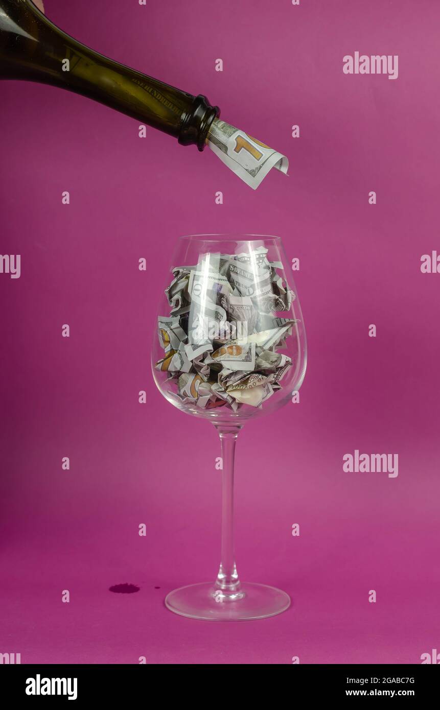 Wine glass is filled with money from bottle. WineGlass is full Crumpled hundred dollar bills on pink background. Stock Photo