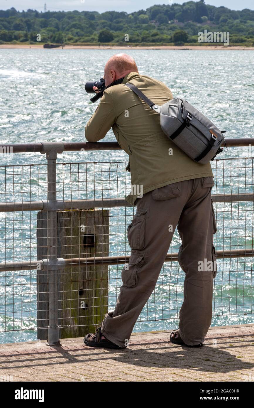 older photographer leaning on railings taking a picture of the sea with a digital camera and holding a camera bag Stock Photo