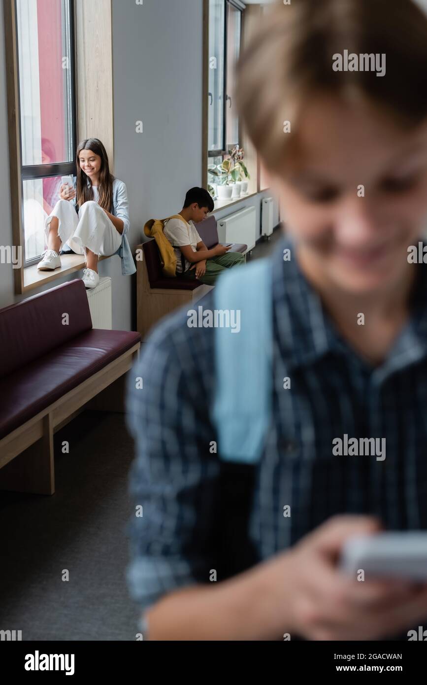 blurred teenager with smartphone near classmates using gadgets on windowsill and bench in corridor Stock Photo