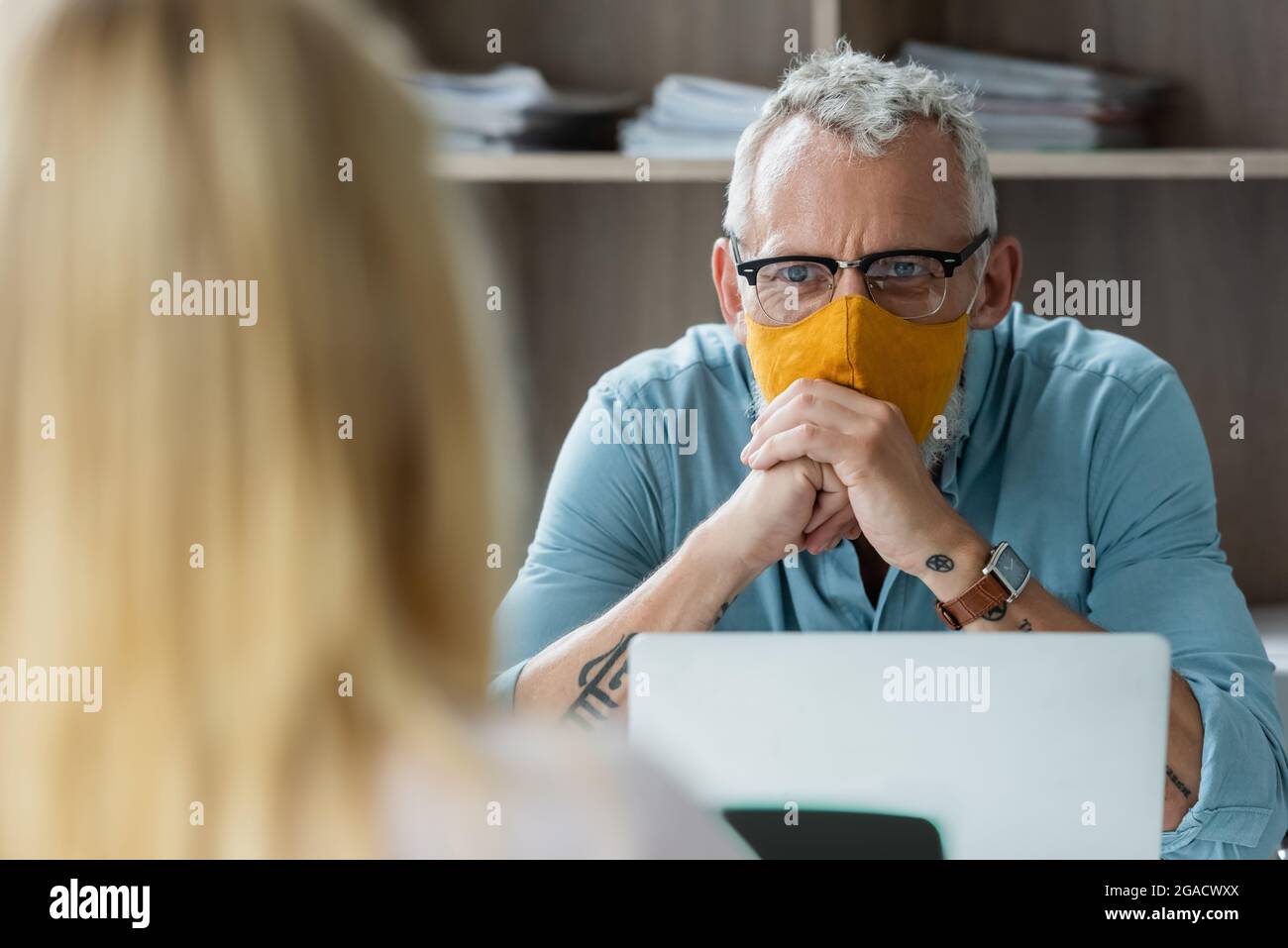 Middle aged teacher in protective mask looking at blurred pupil near laptop in classroom Stock Photo