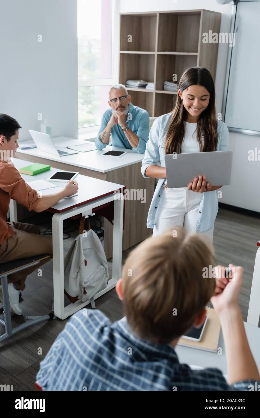 Smiling schoolgirl using laptop near teacher and schoolboys Stock Photo ...