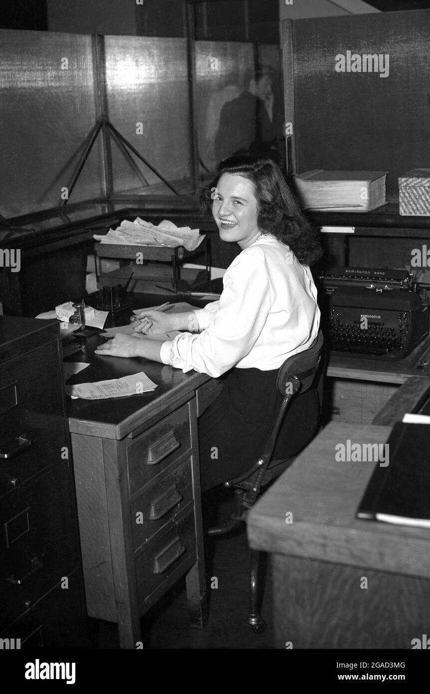 1940s, historical, with a bg smile on her face as she turns for her photo a a young female clerk or secretary sitting at a desk, beside a Remington Rand typewriter, USA. Stock Photo