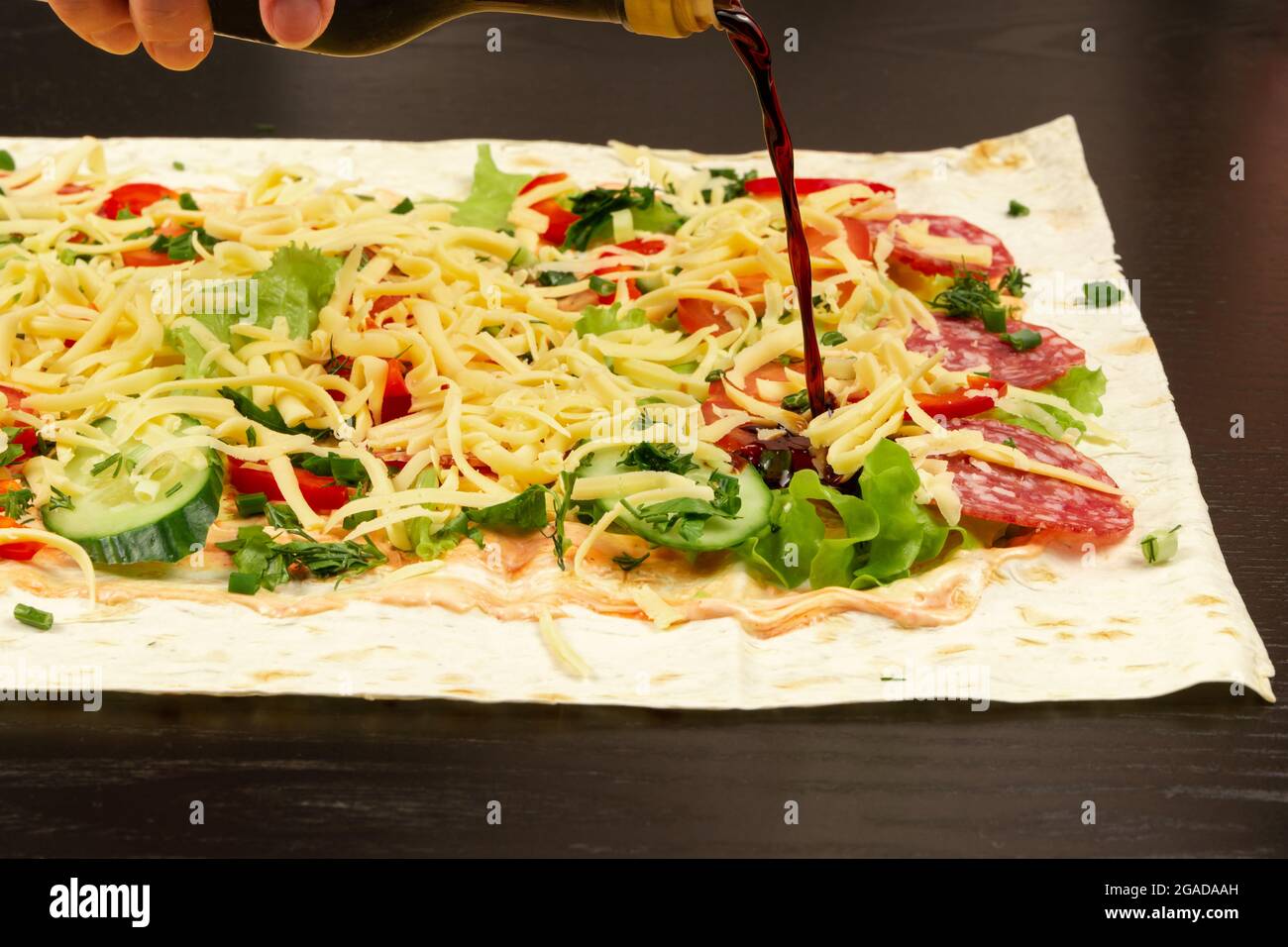 soy sauce is poured from the bottle on the unwrapped shawarma with fresh herbs, cheese and vegetables on the background of a black wooden table Stock Photo