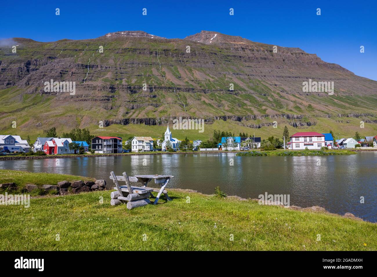 a rustic table and chair overlook the pretty harbour town of seydisfjordur reflected in the calm lagoon on a sunny day Stock Photo