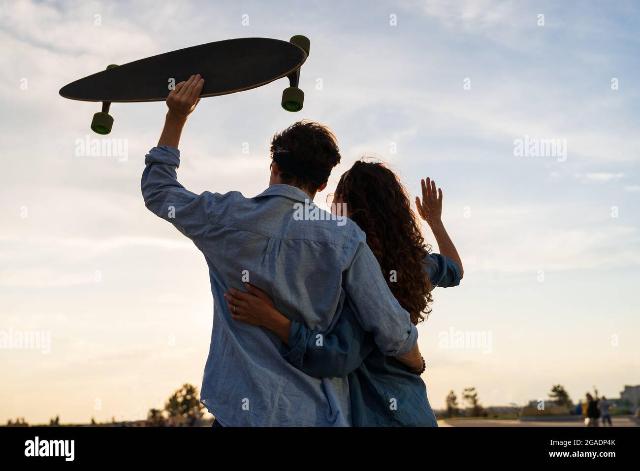 Stylish young couple hug looking at sunset hold skateboard. Happy girl and guy in love enjoy freedom Stock Photo
