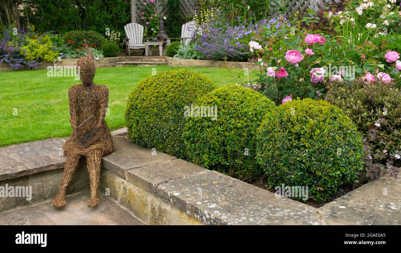 Willow sculpture art (ornamental feature) in beautiful colourful landscaped garden (flowering plants, box balls, lawn, chairs) - Yorkshire England UK. Stock Photo