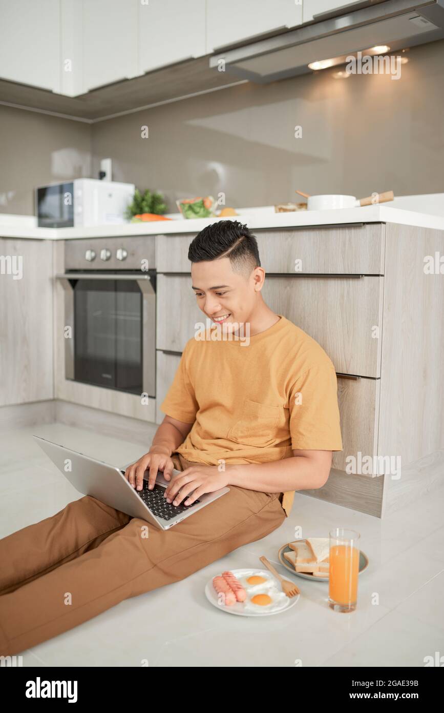 Handsome male sitting on the floor and holding laptop on knees with his breakfast. Stock Photo