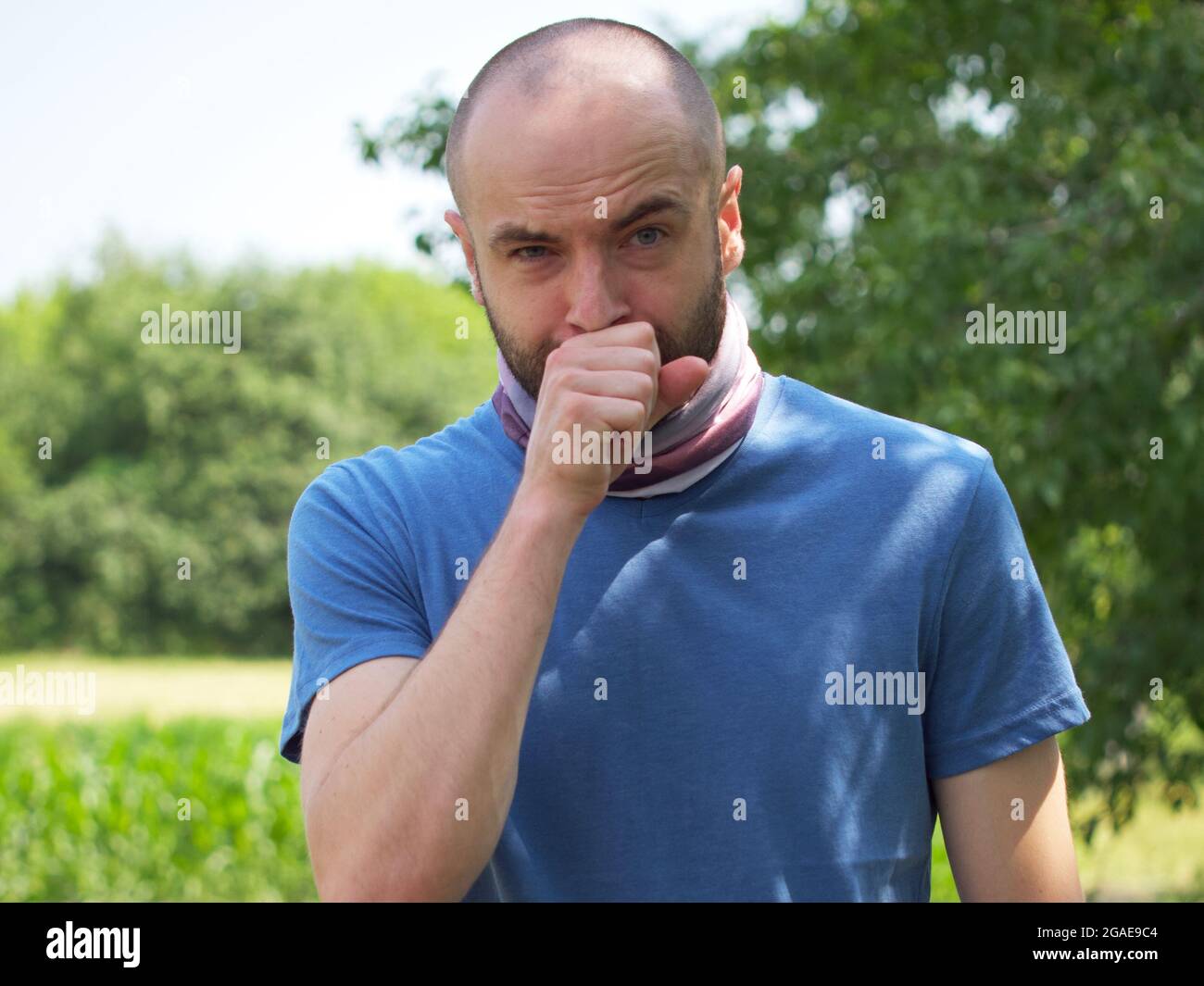 A bald man in a blue T-shirt coughs into a fist. Stock Photo